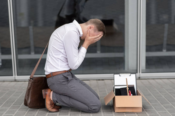 De ontslagen kantoormedewerker viel op zijn knieën en bedekte zijn gezicht door stress. Voor hem staat een kartonnen doos met briefpapier. De man is ongelukkig door een vermindering van de robot. - Foto, afbeelding