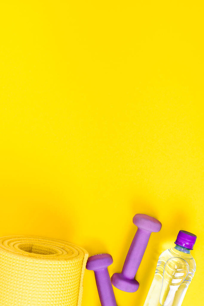 Home fitness concept yellow yoga mat, two purple dumbbells and a bottle of water. Equipment for training and exercises for weight loss. Flat lay a bright vertically background with copy space. - Foto, afbeelding