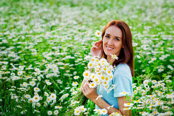 カモミール畑の青いドレスの少女。女の子はたくさんのデイジーと笑顔を抱いています。彼女の髪に花を入れる。草原の夏の自然. - 写真・画像