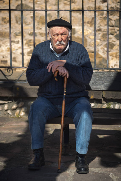 Potes, Cantabria, Spain; 18 03 2017: Elderly man in blue pullover and pants, black beret and club sunbathes sitting on a bench - Foto, Imagen