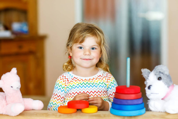Cute little toddler girl playing alone with colorful wooden rainbow pyramid and toys at home or nursery. Happy healthy child having fun in kindergarten or preschool daycare - Foto, Bild