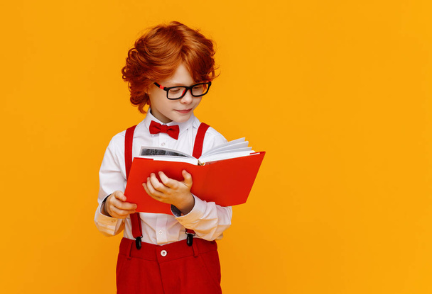 Boldog vörös hajú iskolás fiú szemüvegben mosolyog, miközben könyvet olvas az iskolai tanulmányok során a sárga backgroun - Fotó, kép