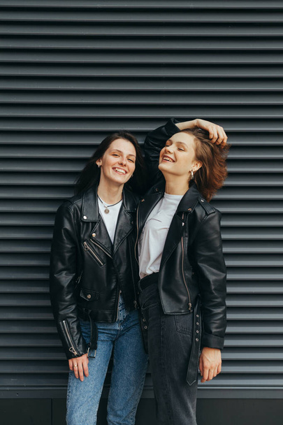 Vertikální fotografie dvou šťastných dívek ve stylovém oblečení stojících na černém pozadí a usmívajících se. Pozitivní dívky v kožených bundách chatování a baví proti černé zdi pozadí. - Fotografie, Obrázek