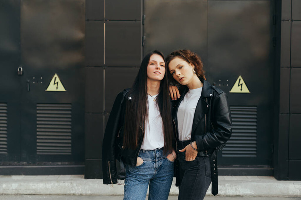 Portrait of two stylish girls in casual clothes posing at camera on black background, wearing leather jackets and jeans. 2 girlfriends in stylish casual clothes on a dark wall background. - Foto, Imagen
