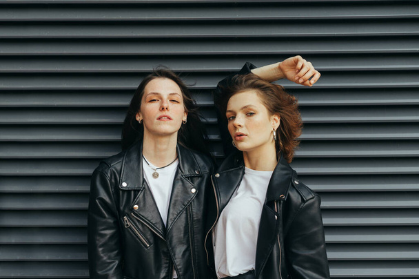 Μοντέρνο πορτρέτο 2 μοντέλων σε κομψά casual ρούχα που ποζάρουν στην κάμερα σε σκούρο φόντο, κοιτάζοντας κάμερα με σοβαρό πρόσωπο.Δύο νεαρές κοπέλες με δερμάτινα μπουφάν στέκονται απέναντι σε έναν μαύρο τοίχο - Φωτογραφία, εικόνα