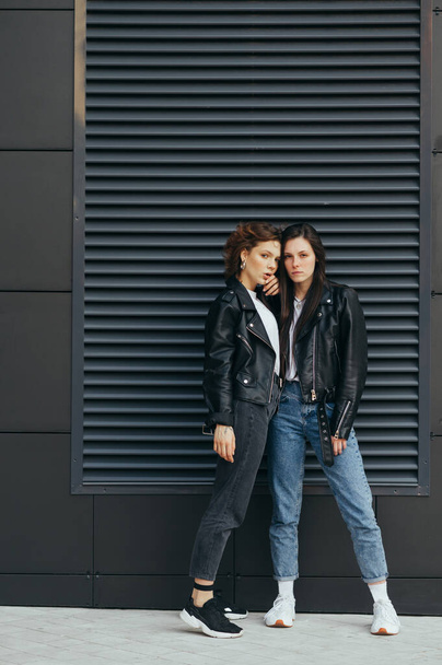 Vertikales Porträt zweier Freundinnen in lässiger Kleidung, in Lederjacken und Jeans, stehend auf schwarzem Hintergrund, posierend vor der Kamera.Modisches Paar von Mädchen posiert an schwarzer Wand - Foto, Bild
