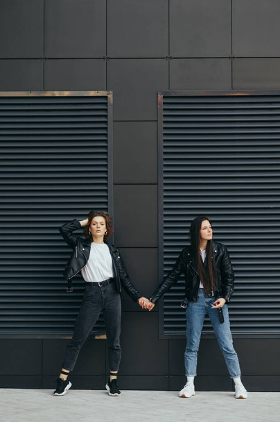 Due ragazze alla moda si tengono per mano contro un muro nero, in posa per la fotocamera. Ritratto di due ragazze attraenti in giacche di pelle in piedi su sfondo nero che si tengono per mano. Foto verticale. - Foto, immagini