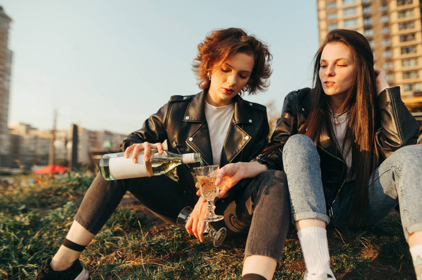 Two attractive girls in stylish clothes sit on the grass against a background of cityscape and pour wine into a glass. Cheerful girls drink wine on a walk in the city, sit on the grass to relax. - Foto, Imagen