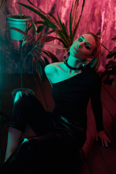 Fashion woman sitting on the floor in tropical foliage neon light. Wet hair, perfect figure and makeup. Pink and green neon color. Beautiful girl and monstera leaves - Foto, Imagem