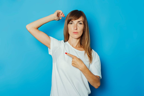 Молодая девушка в белой футболке показывает палец на мокрых подмышках от пота на синем фоне. Концепция чрезмерного потоотделения, жары, дезодоранта
. - Фото, изображение