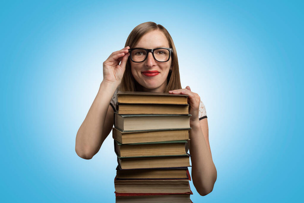 白いドレスと眼鏡の少女が青い背景に本の山に手を置いた。若い学生と教育の概念. - 写真・画像