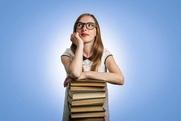 Молода дівчина в білій сукні і сонцезахисні окуляри кладуть руки на купу книг і дивляться на синій фон. Концепція молодого студента та освіти
. - Фото, зображення