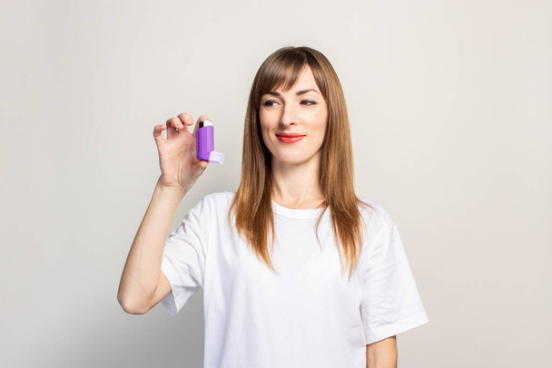 gelukkige jonge vrouw houdt een inhalator in haar hand, kijkt naar de inhalator op een lichte achtergrond. Een spandoek. Concept voor gemakkelijker ademhalen, behandeling van astma, keelholte, strottenhoofd, luchtpijp. - Foto, afbeelding