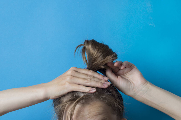 Dívka si narovnává rozcuchaný ocas vlasů na hlavě, na modrém pozadí. Moderní rychlý účes. Vlasy svázané elastickým proužkem. - Fotografie, Obrázek