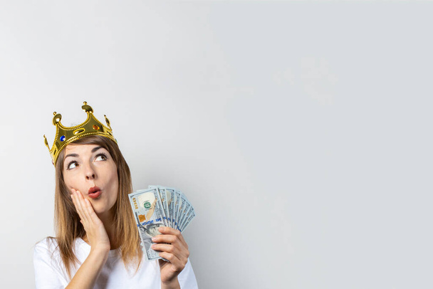 Eine junge Frau mit Krone auf dem Kopf und überraschtem Gesicht hält einen Stapel Geld auf hellem Hintergrund. Emotionales Lachen, Überraschung, Schock. Konzept einer Königin, Glück, Reichtum, Gewinn, Sieg, Wette. Banner. - Foto, Bild