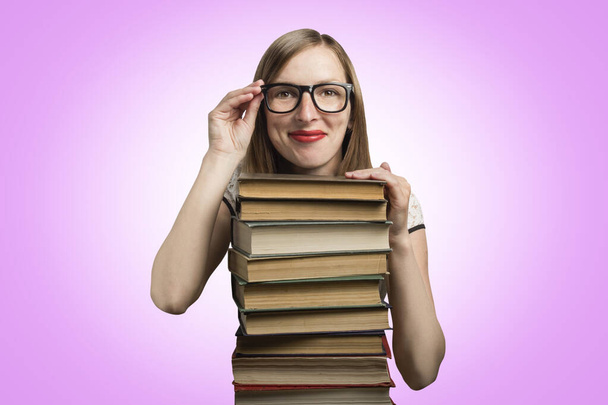 Young girl in a white dress and glasses put her hands on a stack of books on an pink background. Concept of a young student, student and education. - Photo, image
