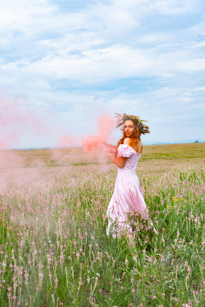 Κοκκινομάλλα κοπέλα σε ένα χωράφι με ένα στεφάνι στο κεφάλι της, κρατά μια έγχρωμη βόμβα καπνού στο χέρι της - Φωτογραφία, εικόνα