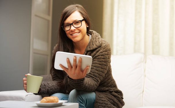 nainen olohuoneessaan ottaa aamiaisen tabletin muki ja brioche, lukea uutisia ja aloittaa työnsä kotoa - Valokuva, kuva