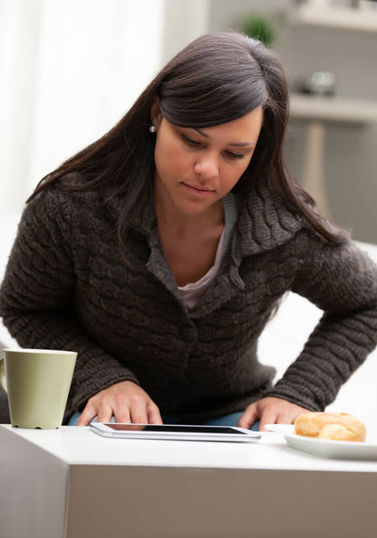 mulher em sua sala de estar tomando café da manhã com caneca tablet e brioche, lendo notícias e começando seu trabalho a partir de casa
 - Foto, Imagem