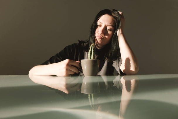 Diet concept: young upset woman in a gray t-shirt holds in her hands a coffee cup with a cactus. The fight against excess weight and extra pounds. - Foto, Imagem