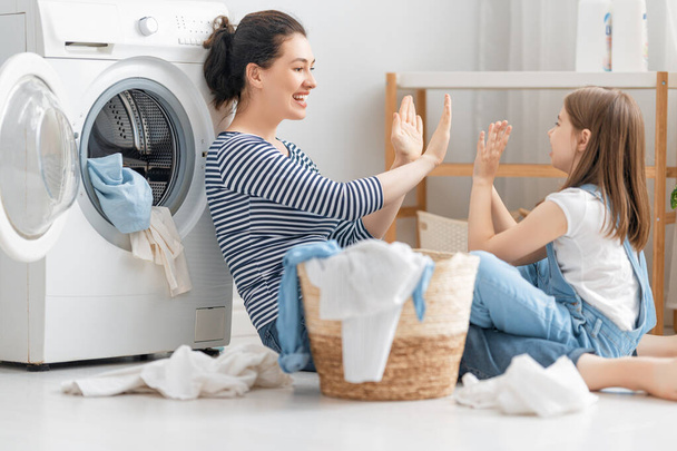Hermosa mujer joven y niña pequeña ayudante se divierten y sonríen mientras lavan la ropa en casa. - Foto, Imagen
