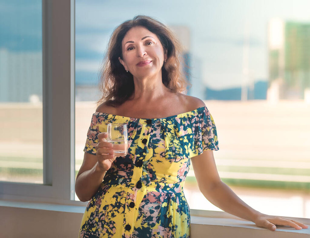 Indoor portrait of a beautiful smiling senior woman next to window holding glass of water  - Photo, Image