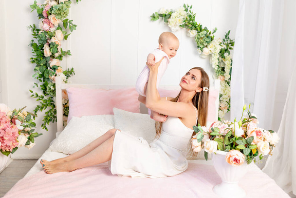 a young beautiful mother with a 6-month-old daughter in her arms sits on a white bed in flowers and hugs her, a place for text - Photo, Image