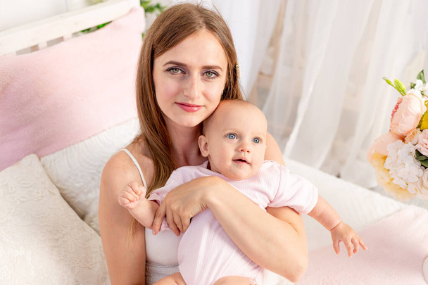 uma jovem mãe bonita com uma filha de 6 meses de idade em seus braços senta-se em uma cama branca em flores e a abraça, um lugar para texto
 - Foto, Imagem