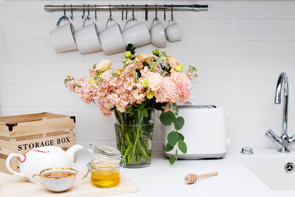 wedding bouquet of pink and orange lisianthus, antirrhinum and eucalyptus in glass vase on kitchen table. Black tea in a white cup, honey jar and spoon for honey, lemon closeup on a white kitchen - Foto, afbeelding