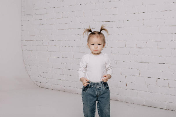 cute little girl with a ponytail hairstyle dressed in a white T-shirt and denim pants posing for a photo on a white background in a photo studio - Fotó, kép