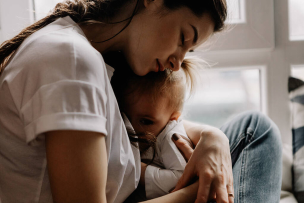 young pretty woman breastfeeding her little daughter sitting on the window sill of her bedroom - Фото, изображение