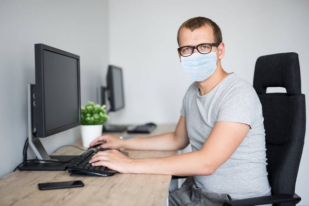 coronavirus, pandemic, quarantine, health care and office work concept - portrait of young handsome man in protective mask using computer in office or at home - Photo, Image
