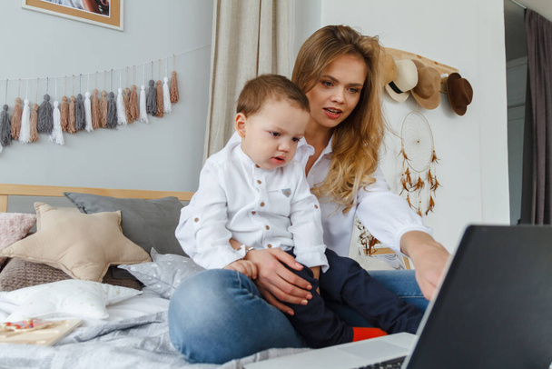 A young mother with her beloved son in the bedroom on the bed with a laptop. Work at home with a child concept - Foto, Imagem