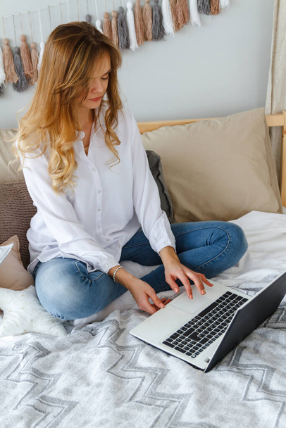 Young beautiful girl with a laptop in the bedroom sits on the bed - Foto, Bild