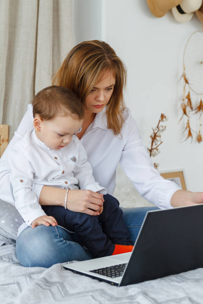 A young mother with her beloved son in the bedroom on the bed with a laptop. Work at home with a child concept - Foto, Imagem
