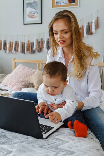 A young mother with her beloved son in the bedroom on the bed with a laptop. Work at home with a child concept - Foto, imagen