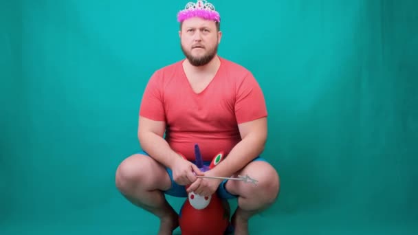handsome bearded freaky man in a pink T-shirt with a deadema on his head is saddened riding a unicorn with a magic wand in his hand. A funny wizard joke to make and fulfill a wish. - Footage, Video