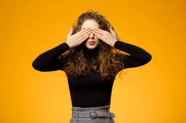 Pretty girl hide her face with hands isolated on yellow background. Model with long brown curly hair - Photo, Image
