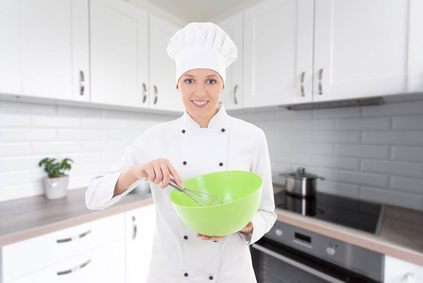 hermosa mujer en uniforme de chef mezclando algo en un tazón de plástico verde en la cocina moderna
 - Foto, Imagen