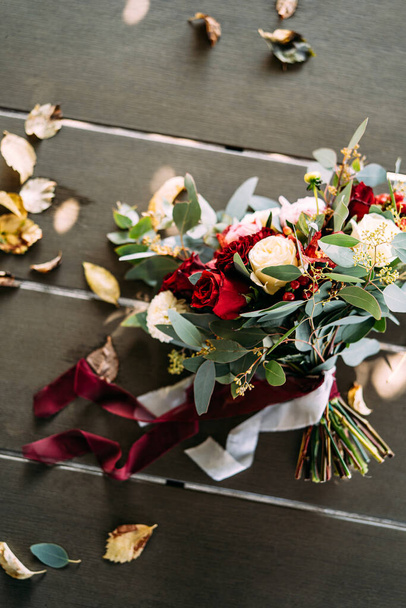 menyasszonyi csokor fehér és vörös rózsából, bazsarózsából, eukaliptikus fa ágaiból, alstroemériából és krizantémból, vörös bogyók fehér és barna szalaggal az asztalon  - Fotó, kép