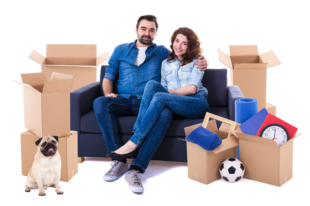 moving day and new home concept - lovely couple sitting on sofa with brown cardboard boxes and dog isolated on white background - Photo, Image