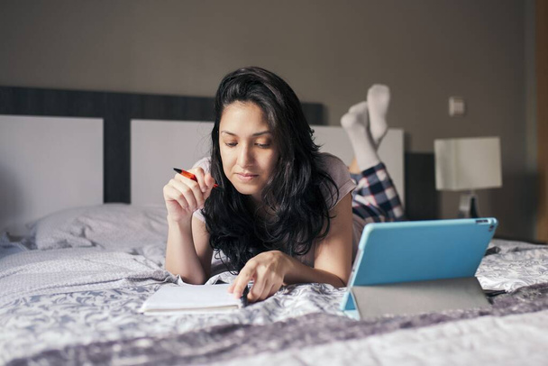 Een vrouw die in een notitieboekje opschrijft, online studeert met een tablet die thuis op bed ligt - Foto, afbeelding