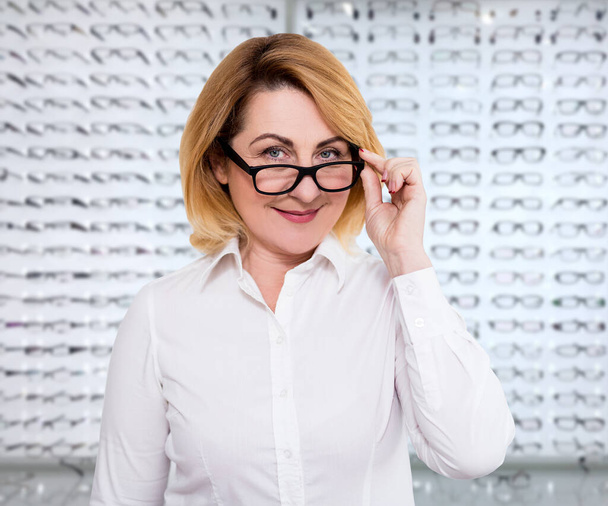Seh- und Sehkonzept - Porträt einer lächelnden reifen Frau beim Ausprobieren einer neuen Brille im Optikergeschäft - Foto, Bild