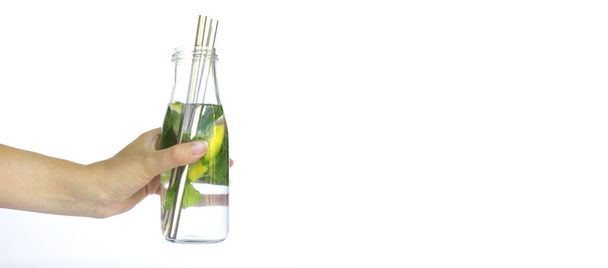 Cold drink with lemon and mint in a transparent glass bottle on a hot summer day. Woman hand holding a bottle with metal eco cocktail straw on it on white background with space for text. Isolated - Фото, изображение