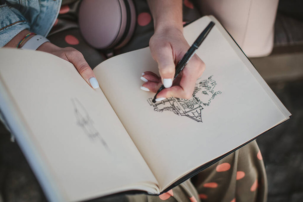 Скетчбук с рисунком дома крупным планом. Девушка рисует левой рукой
 - Фото, изображение