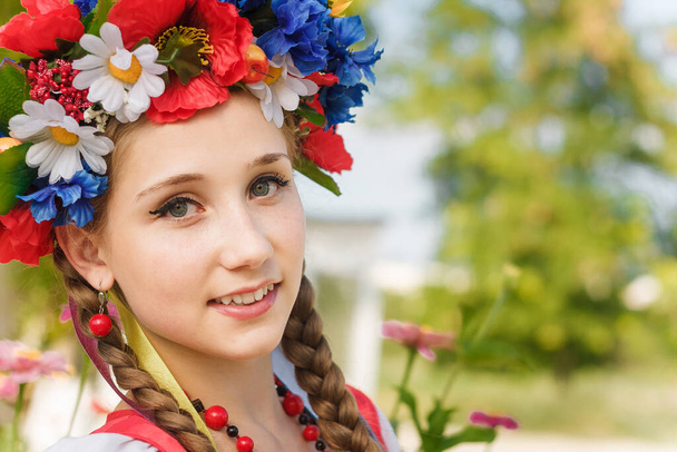 Близкий портрет девушки с двумя косичками в украинских традиционных цветочных венках с лентами, красными серьгами и бусами. Заголовок на размытом внешнем фоне, пространство для копирования
.  - Фото, изображение
