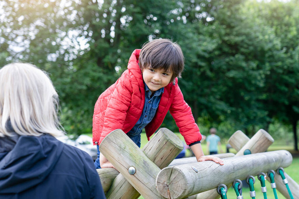 Portret kind klimmen op houten roem in het park met wazige voorgrond van oma, Kind genieten van de activiteit in een klimpark op zomerse zonnige dag, kleine jongen plezier in de speeltuin - Foto, afbeelding