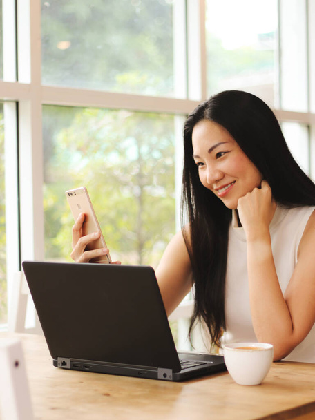 Όμορφη Ασιάτισσα επιχειρηματίας με μακριά μαύρα μαλλιά κάθεται στην καφετέρια με τον υπολογιστή στο τραπέζι χαμογελώντας και κάνοντας τη δική της φωτογραφία με το κινητό τηλέφωνο. - Φωτογραφία, εικόνα