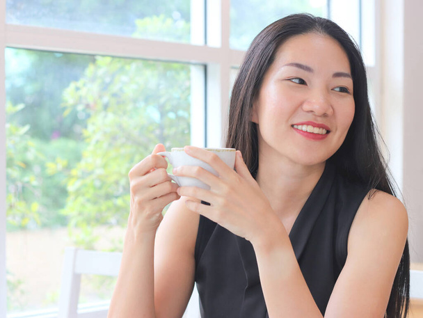 Kaunis aasialainen nainen pitkät mustat hiukset tilalla kuppi kahvia istuu ikkunan kahvilan hymyillen iloisesti ja sivuun. - Valokuva, kuva