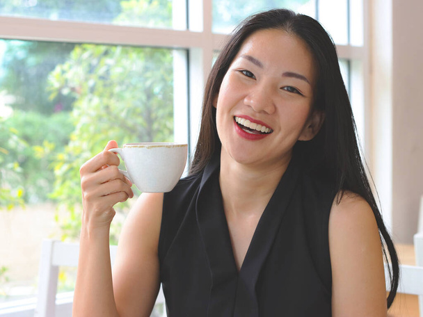 長い黒髪の美しいアジアの女性がコーヒーを飲みながらカフェに座って笑ってカメラを見て. - 写真・画像